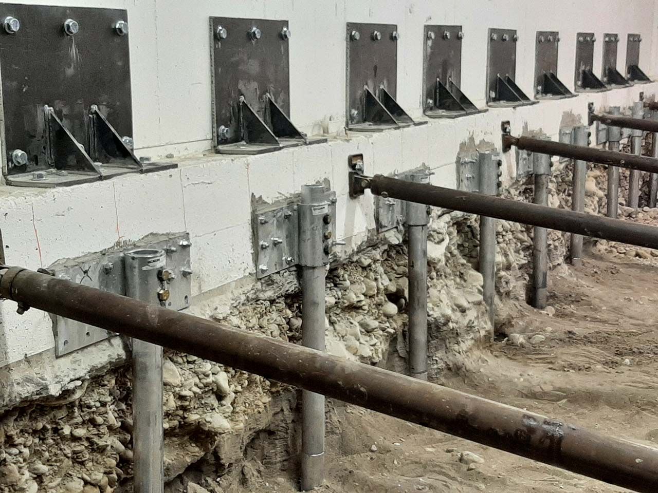 Steel push-pier brackets bolted along a foundation wall, with pier pipes driven into the soil below